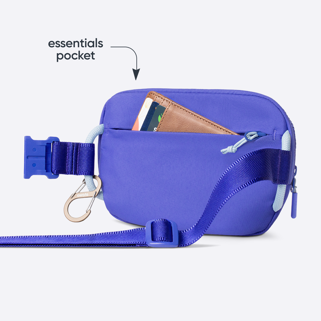 Owala Daily Crossbody in Al Fresco, periwinkle blue belt bag with matching strap, buckle, carabiner, and zipper, shown from the back with a brown leather card wallet tucked into the rear zip pocket, with text reading "essentials pocket"